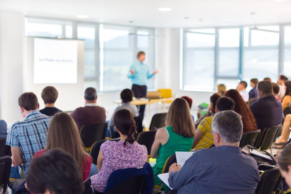 Audience attending a business presentation in a bright conference room, focused on a speaker near a projector screen