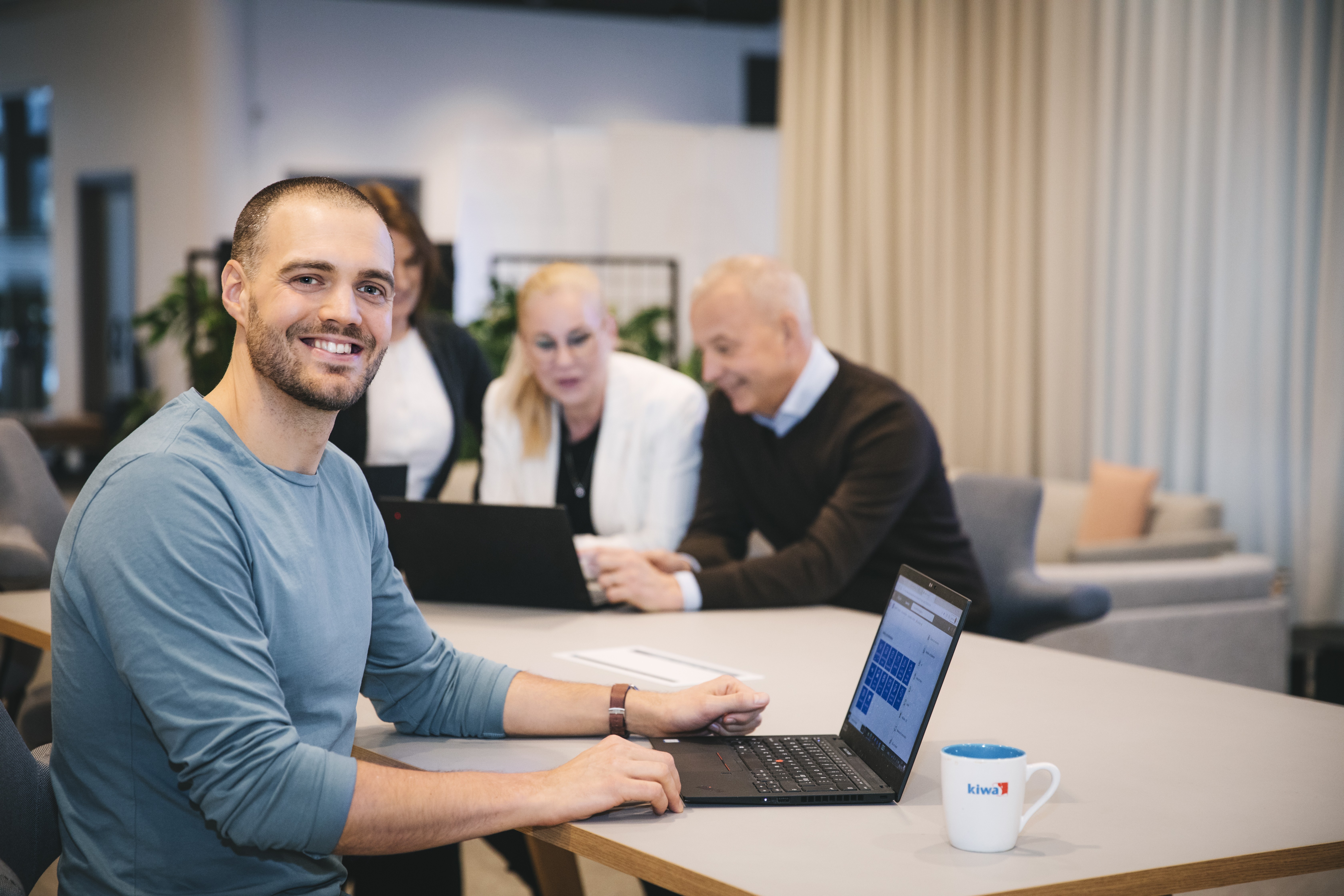 Smiling man in a blue shirt using a laptop at a conference table with coworkers in the background.