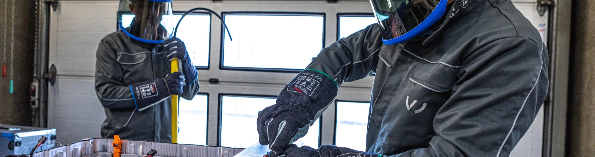 Two workers in protective gear handling equipment in an industrial workshop. Safety face shields, gloves and workwear in a manufacturing setting.