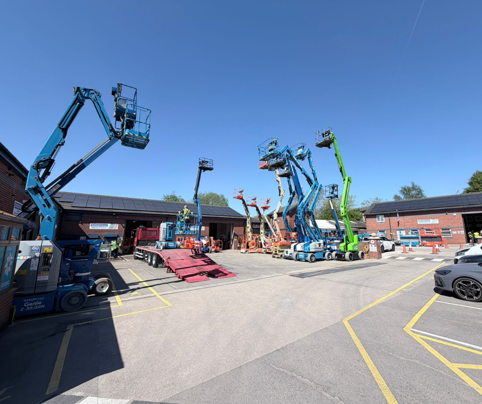 En rad höjdarbetsplattformar och skyliftar som står parkerade på en parkeringsplats utanför industriella tegelbyggnader under en klarblå himmel.