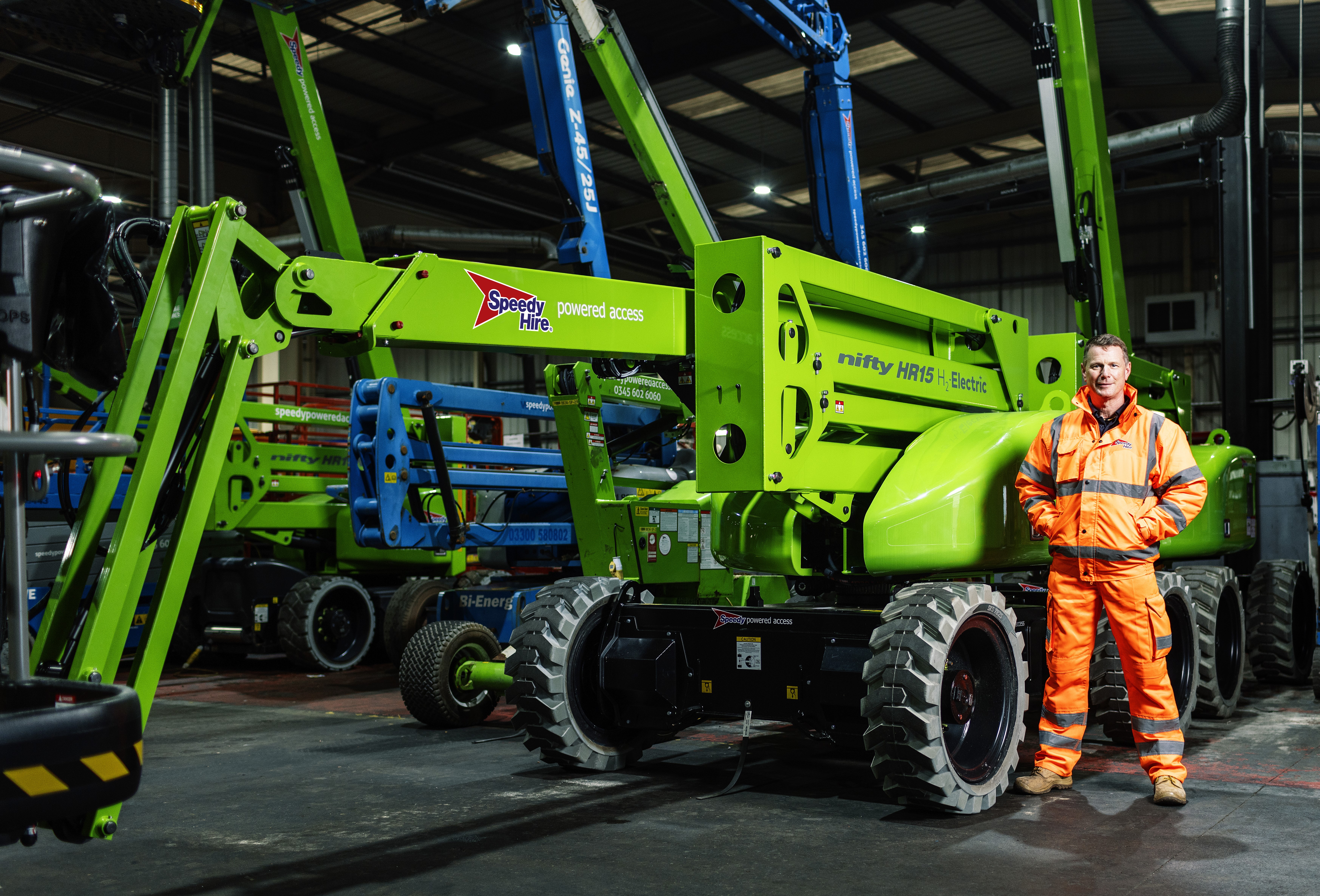 En man i orange varselkläder står bredvid en limegrön Genie HR15 Hybrid, en elektrisk bomlyft, i ett maskinlager inomhus.