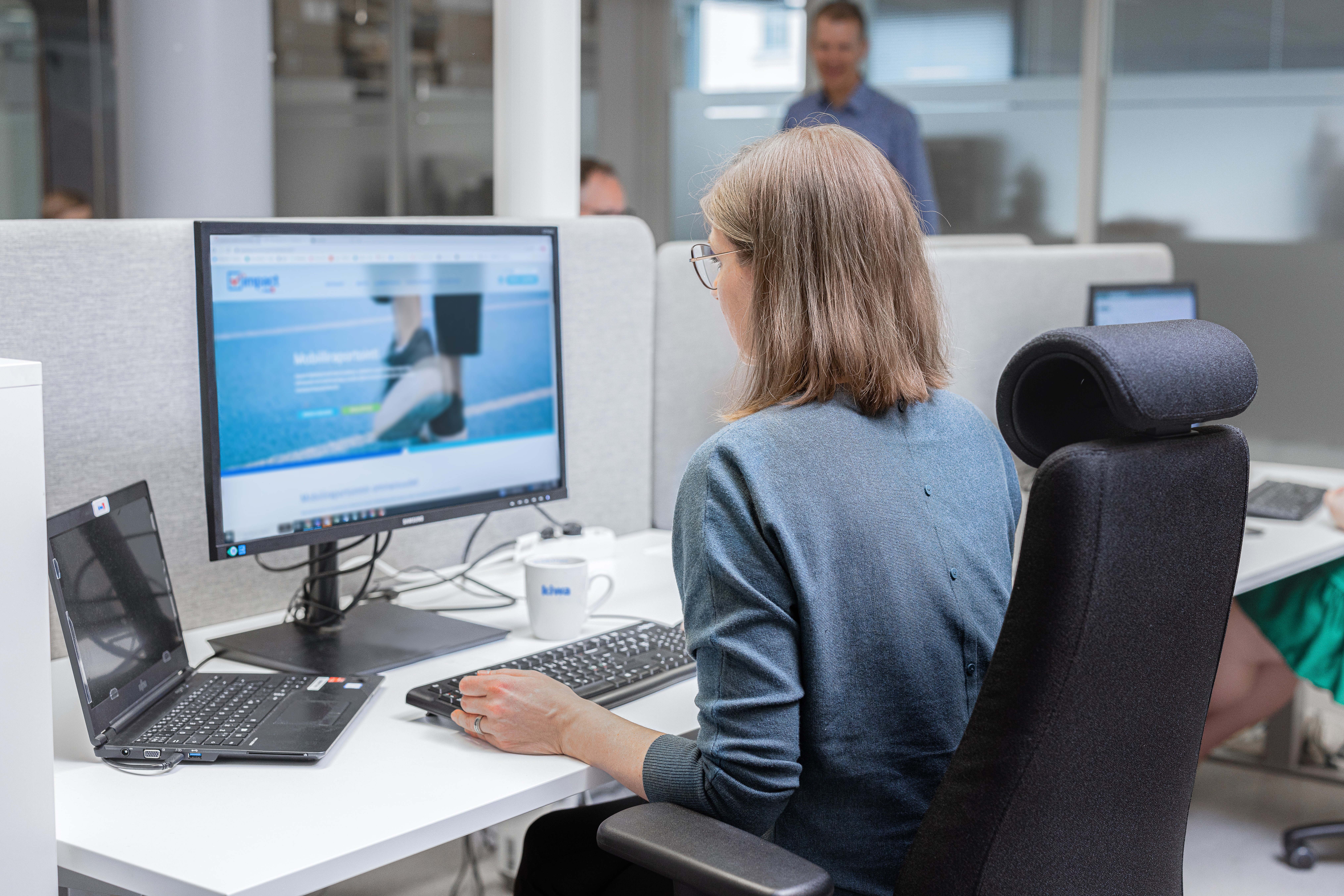 Woman working at a desk with a desktop computer and laptop in an office cubicle.