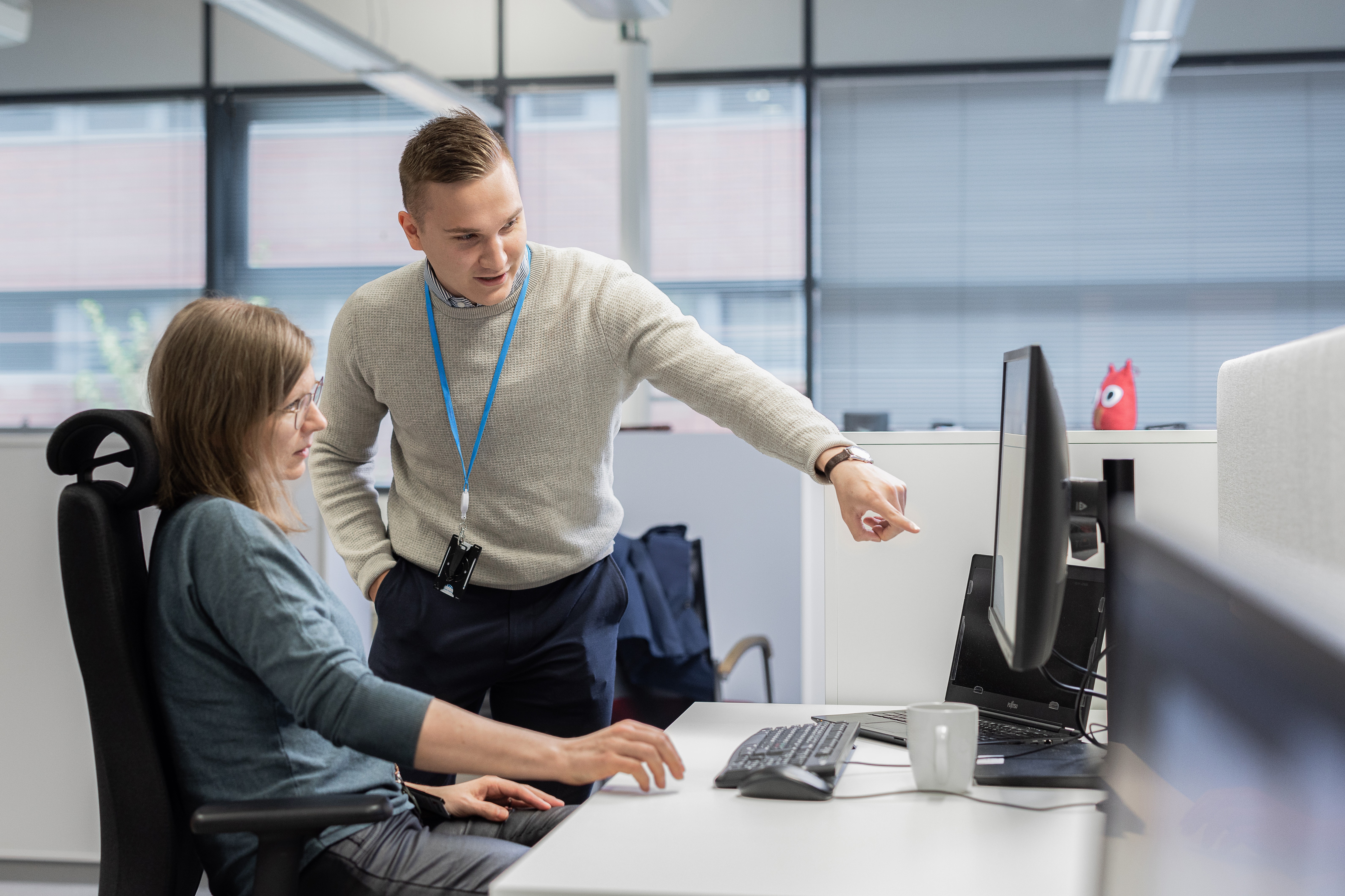 Man pointing at a computer monitor while talking to a woman seated at a desk in an office.