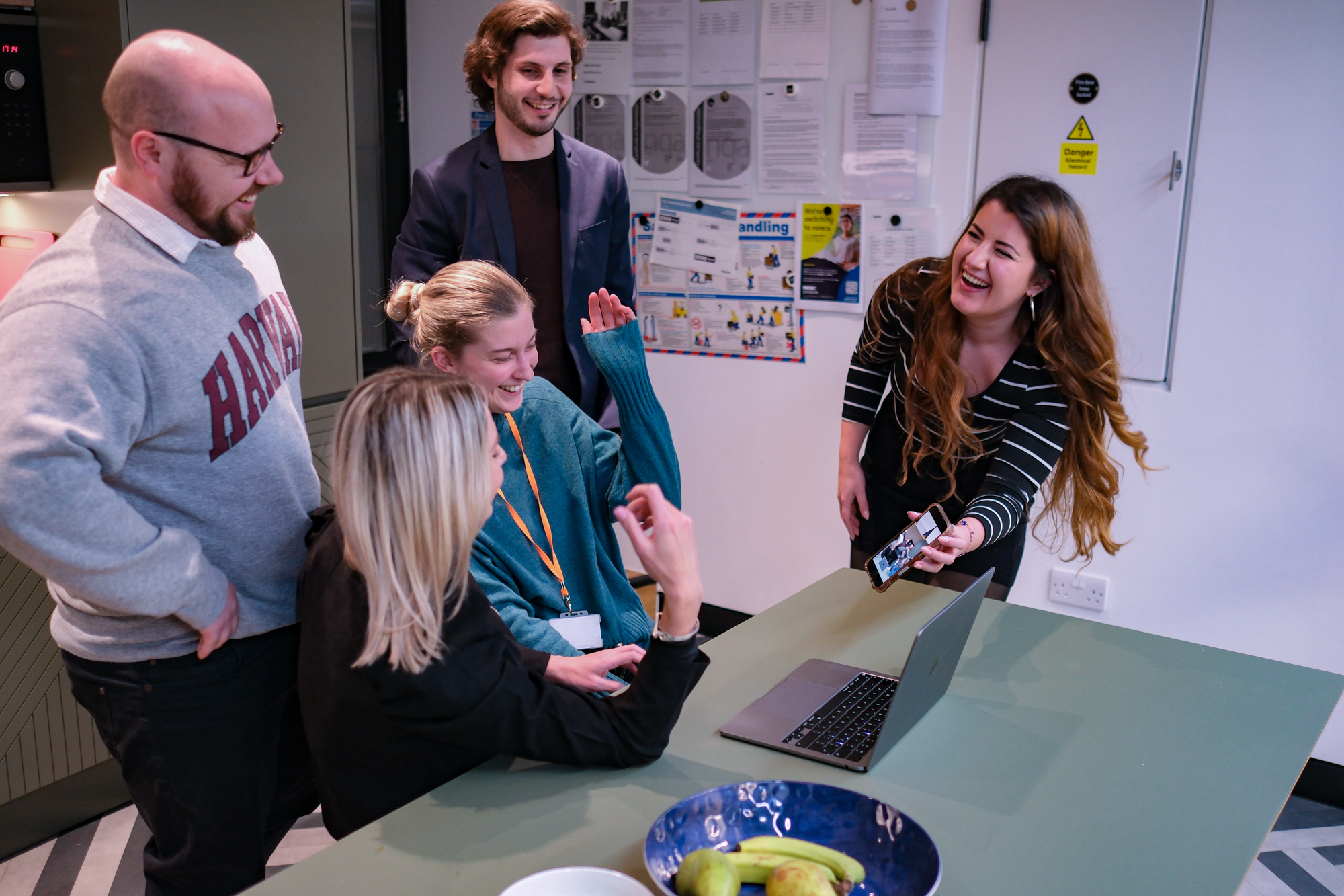 Group of five coworkers gathered around a laptop in an office, smiling and chatting while one woman holds up a smartphone.