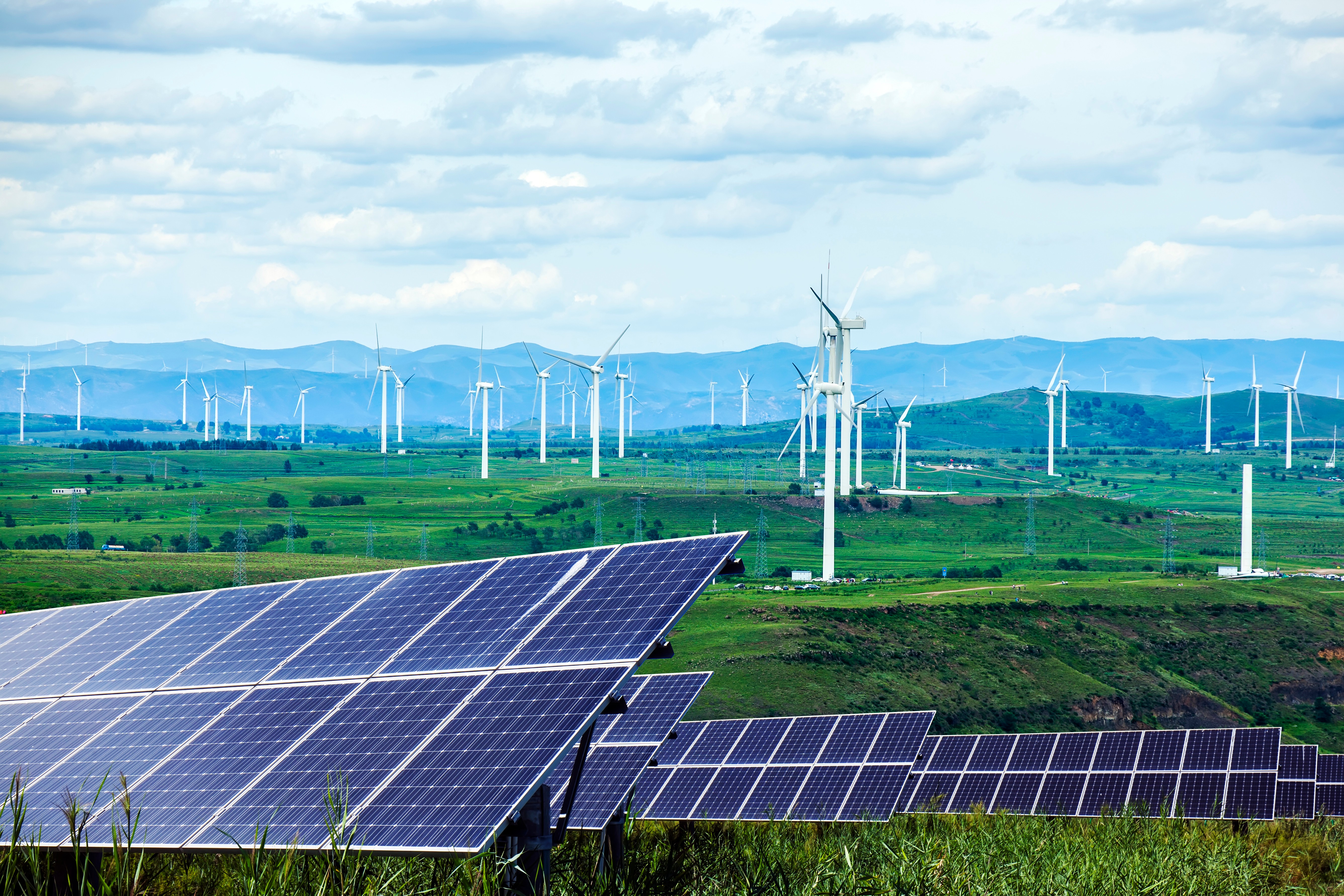 Solar panels and wind turbines in a vast green landscape under a cloudy sky.