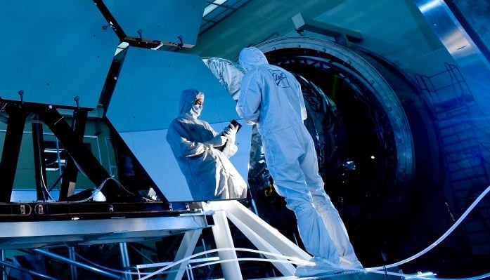 Scientist in protective suits working on a large telescope mirror inside a high-tech laboratory, highlighting precision and innovation in optical technology.