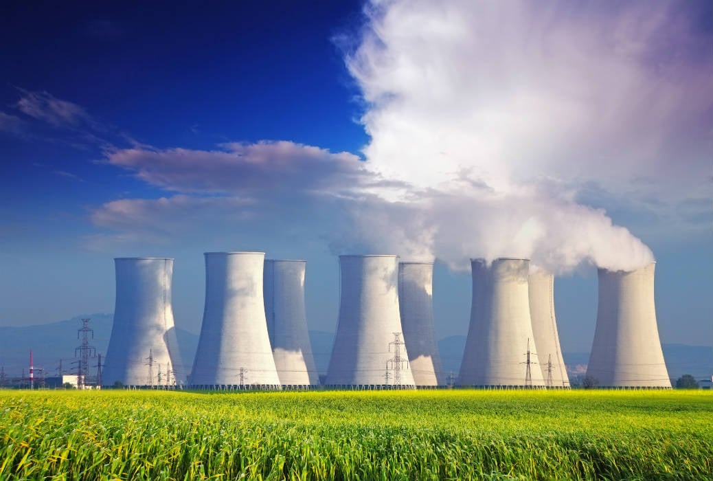 Nuclear power plant cooling towers emitting steam against a blue sky, surrounded by green fields.