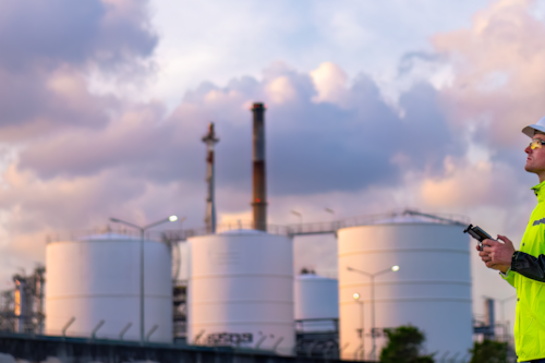 Industrial engineer in high-visibility clothing and hard hat operates equipment at an oil refinery with storage tanks and smokestacks in the background during a cloudy sunset