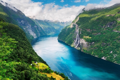 Naturbild visar en fjord mellan gröna höga berg som har snötäckta toppar längst bort i horisonten.
