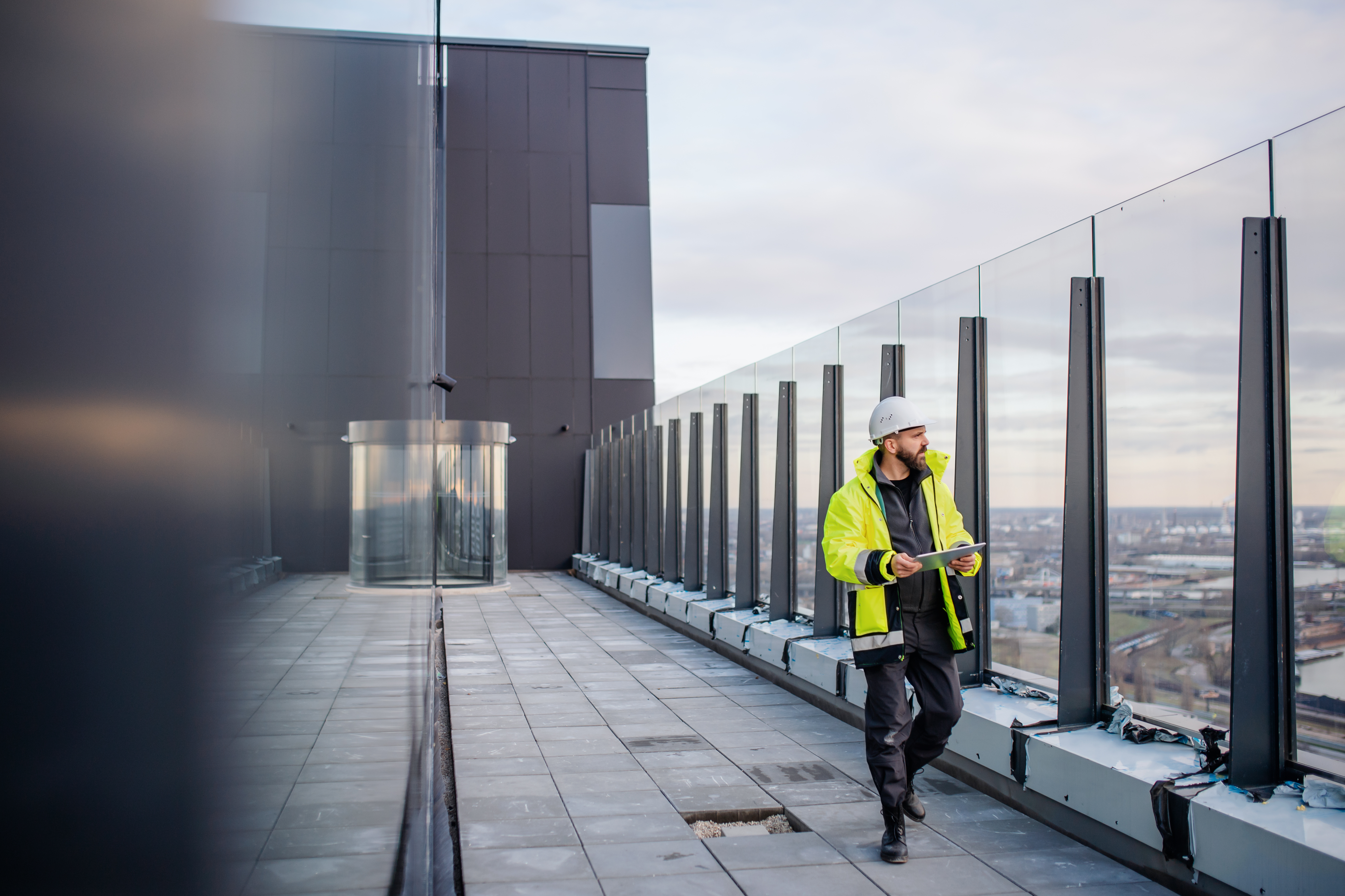 Kiwa inspector inspecting a rooftop with reflective glass, wearing a high-visibility jacket and hard hat, cityscape in the background.