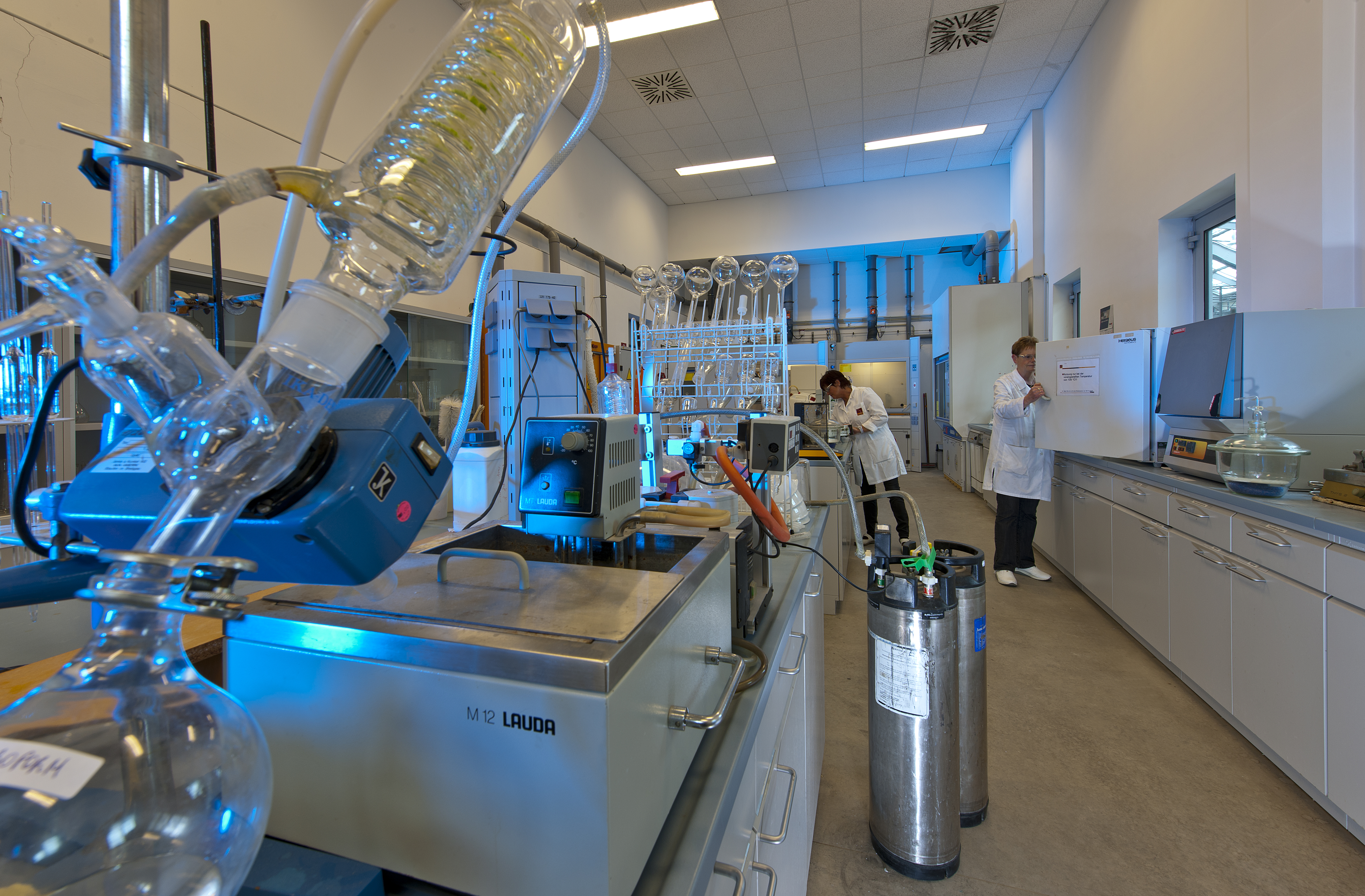 Laboratory with glass distillation equipment, lab benches, and two Kiwa laboratory employees in white lab coats working indoors.