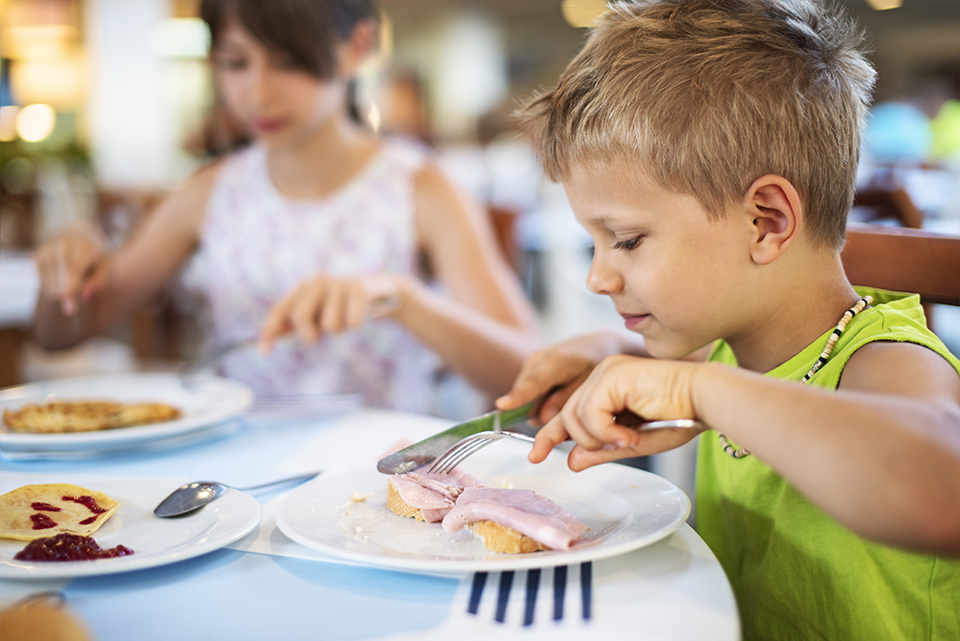 Niño comiendo en un restaurante, con platos de comida como pan y jamón sobre la mesa.
