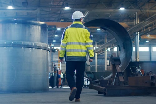 En entreprenadbesiktningsman promenerar bort, klädd i varseljacka och bygghjälm, och ser sig omkring på en konstruktionsplats.