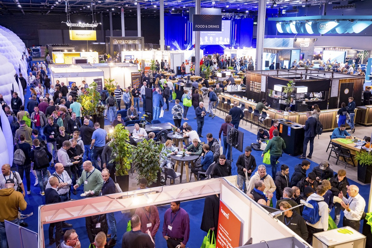 Beursvloer met veel bezoekers, informatiestands, zitjes en een food- en drinkszone in een grote hal.