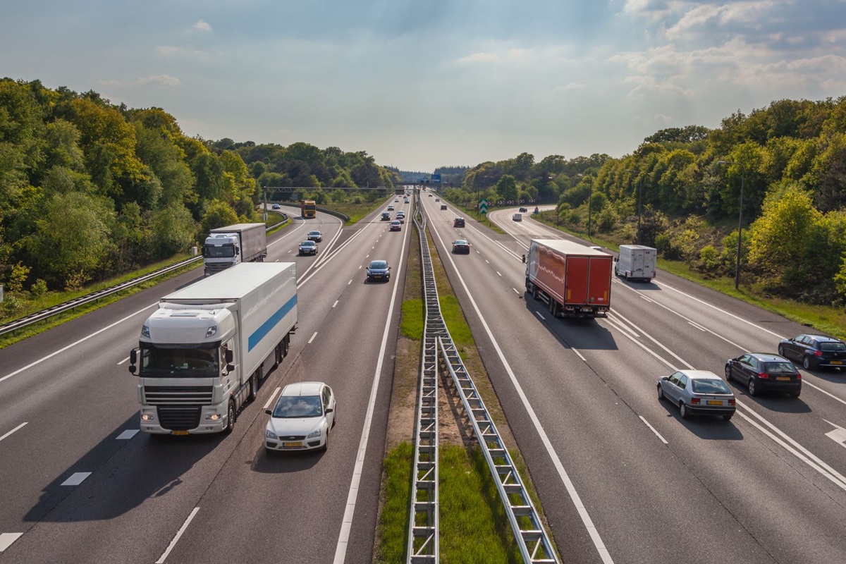 Drukke Nederlandse snelweg met meerdere vrachtwagens en auto’s, gezien vanaf een viaduct, omgeven door groen landschap en onder een licht bewolkte lucht.