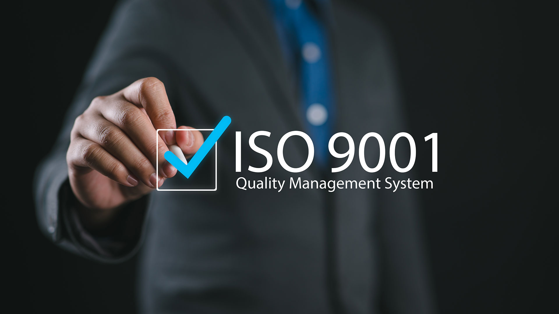 Hand som markerar en kryssruta bredvid texten ISO 9001 och Quality Management System, med en person i mörk kostym i bakgrunden.