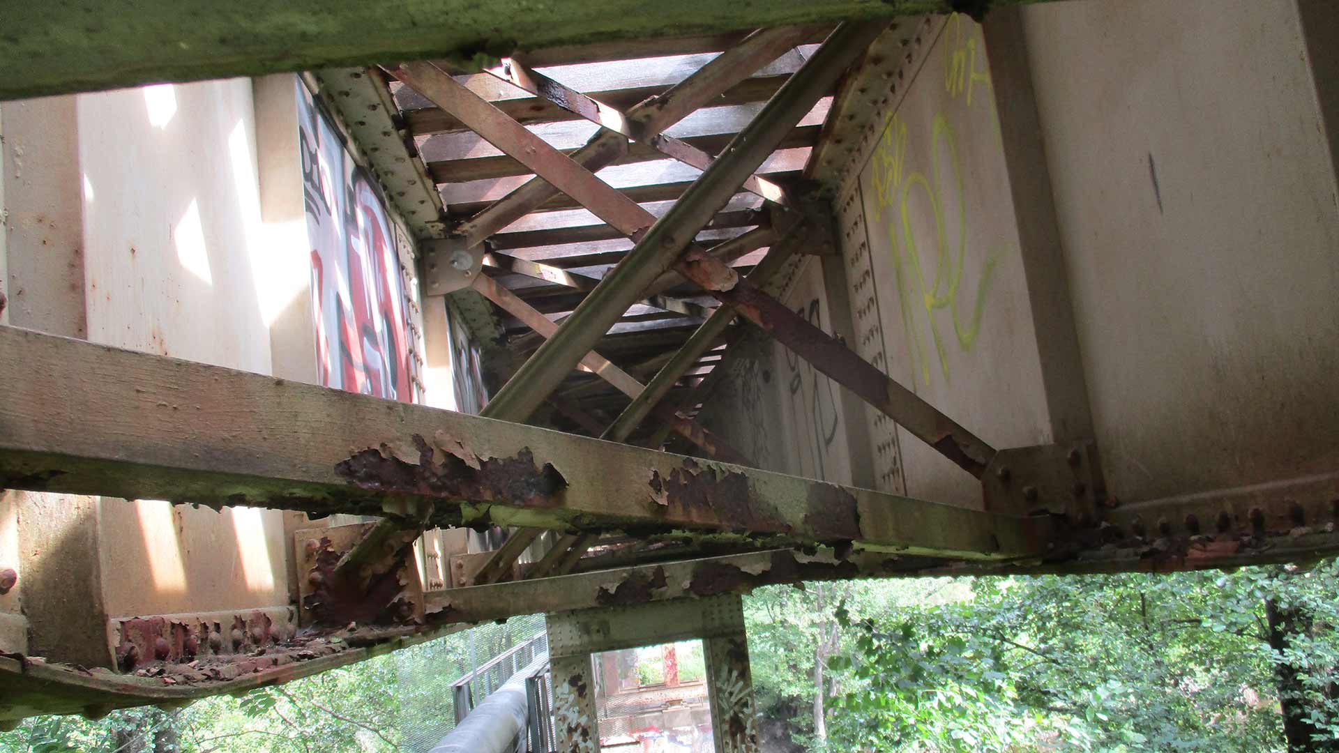 En rostig metallbro med korsande stödkonstruktion, sett från undersidan, med grön vegetation och graffiti på väggen.