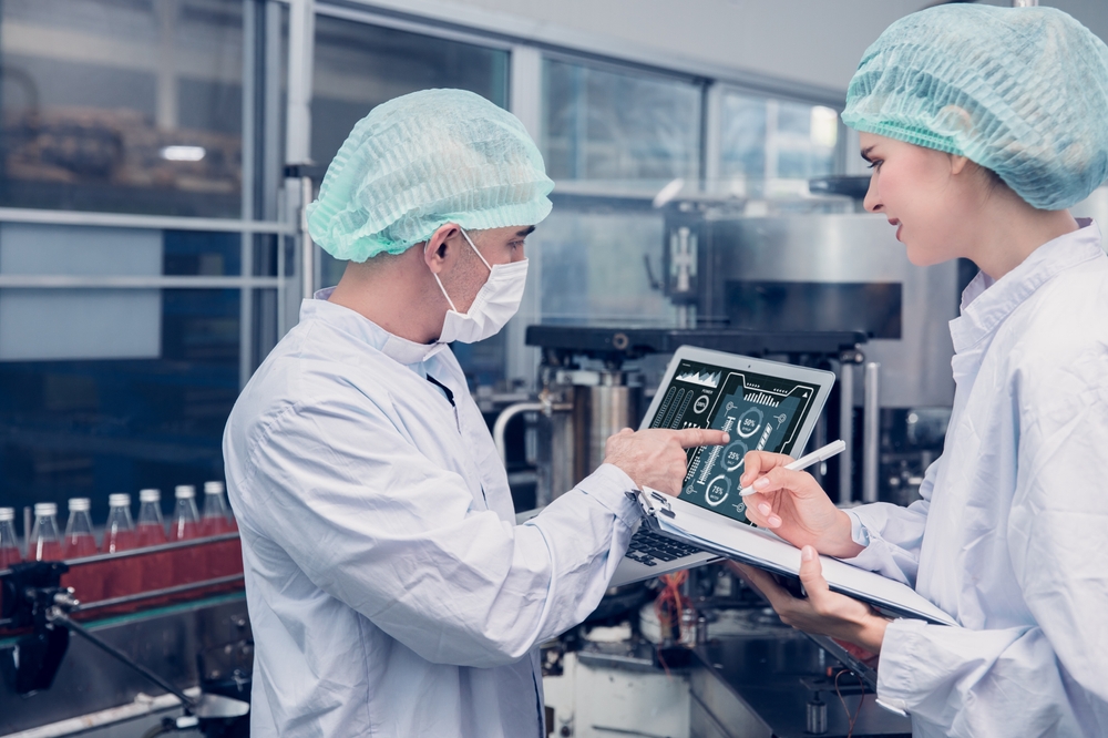 Two Kiwa inspectors in lab coats and hairnets reviewing a digital tablet and clipboard in a manufacturing facility.