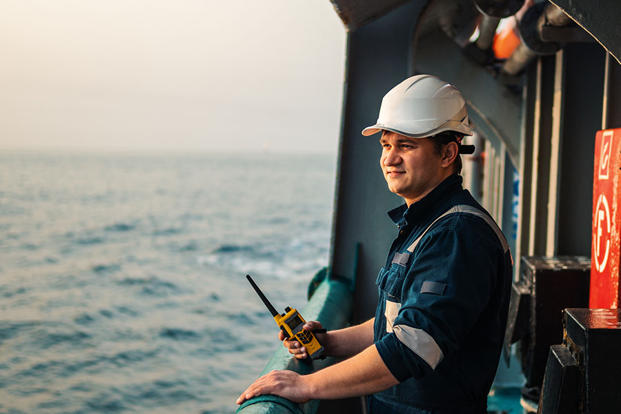 Portrait of a man in a white hard hat holding a walkie-talkie on a ship deck overlooking the ocean.
