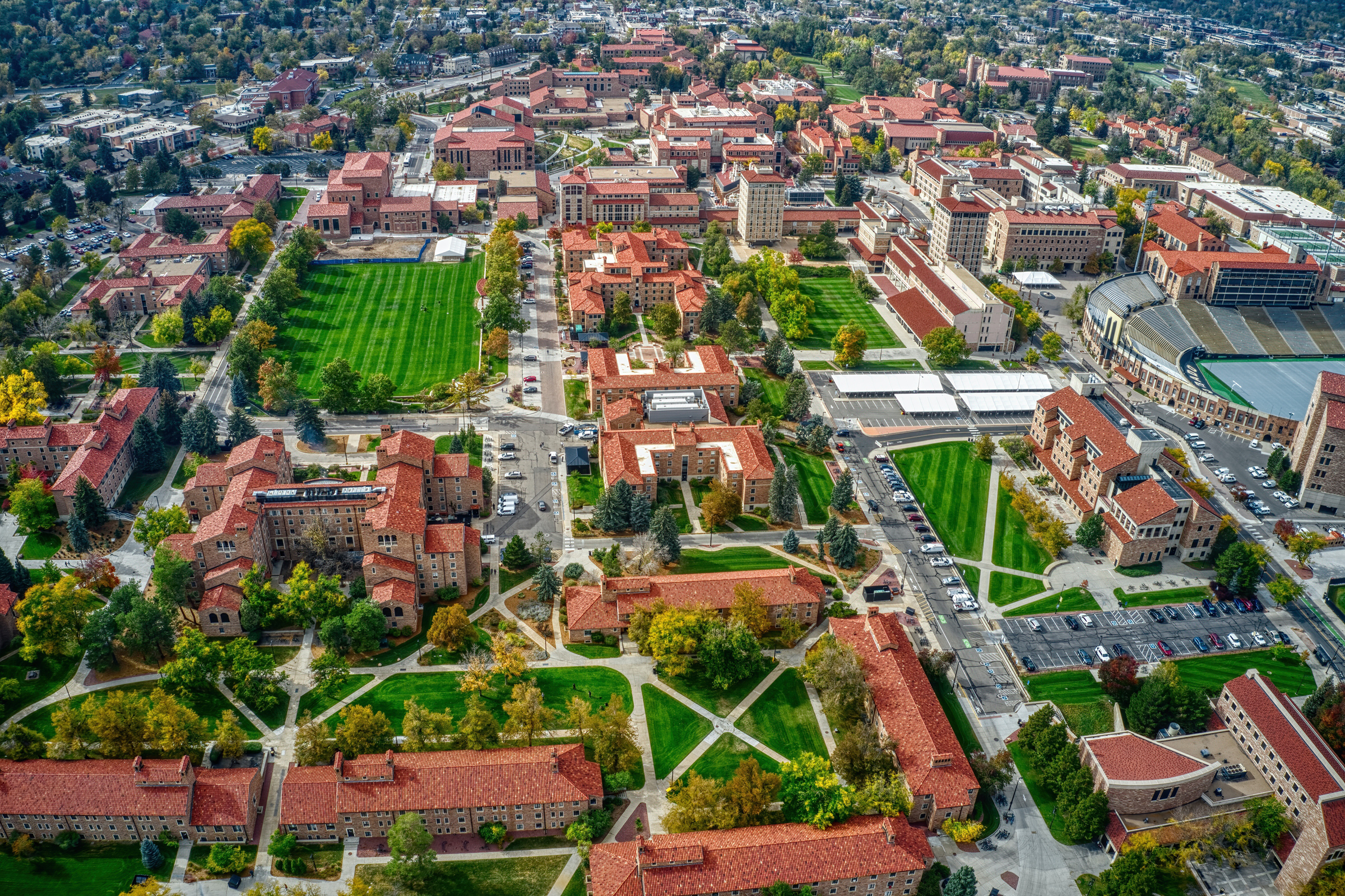Aerial view of a university campus with red-roofed buildings, green lawns, and surrounding trees. Parking areas and a stadium are visible in the background.