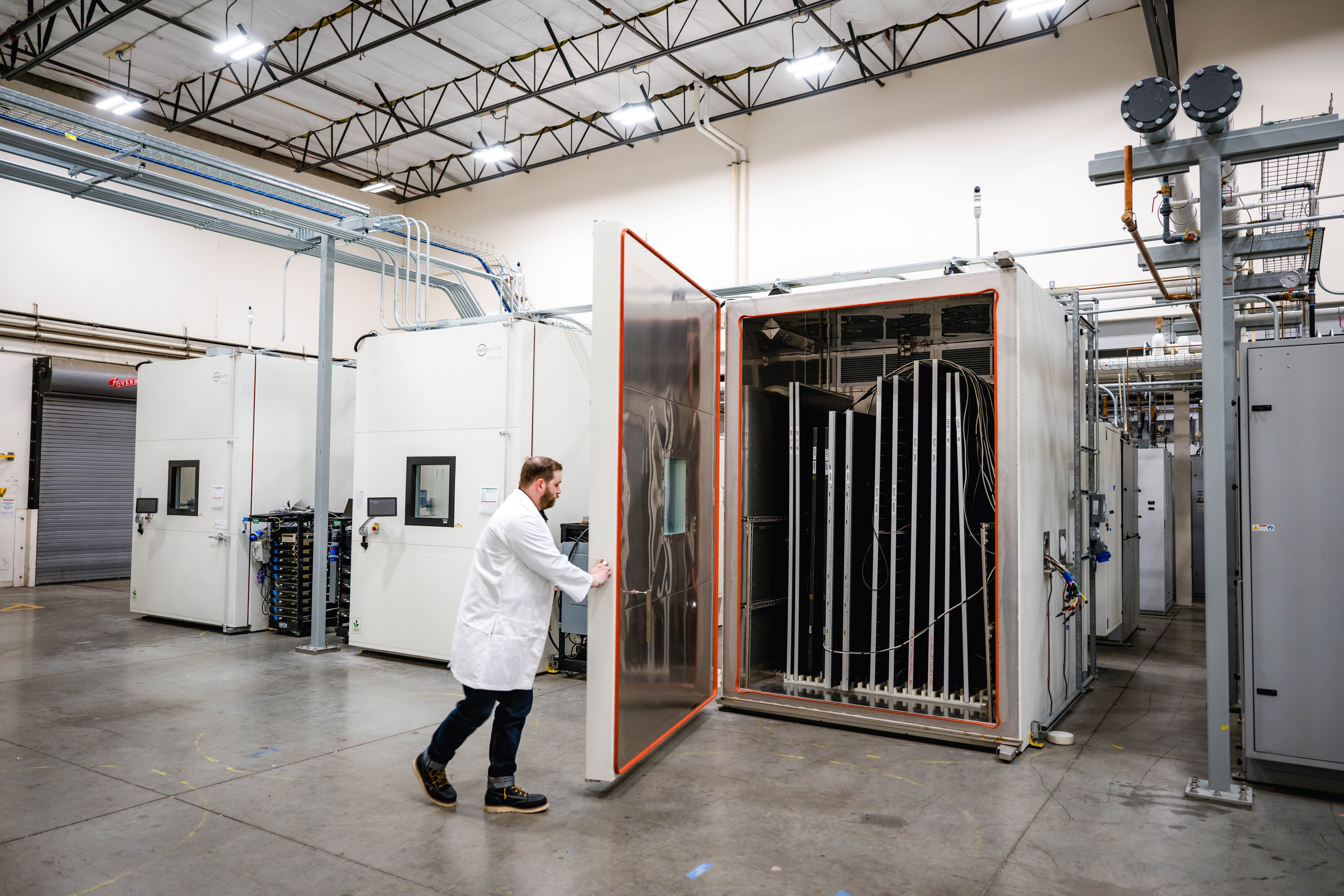 A Kiwa PVEL laboratory employee wearing a white lab coat opens the door to a large industrial testing chamber in a spacious warehouse setting.